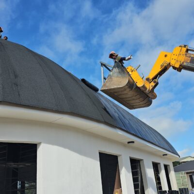 Workers installing roofing materials on a large curved roof structure during a commercial construction project.